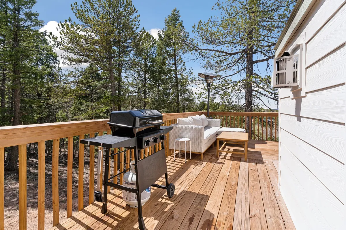 A spacious wooden deck set amidst lush trees, featuring a gas grill, plush outdoor seating, and cozy Adirondack chairs. The inviting setup encourages social gatherings and relaxing moments, with the grill ready for cookouts and ample seating for family and friends.