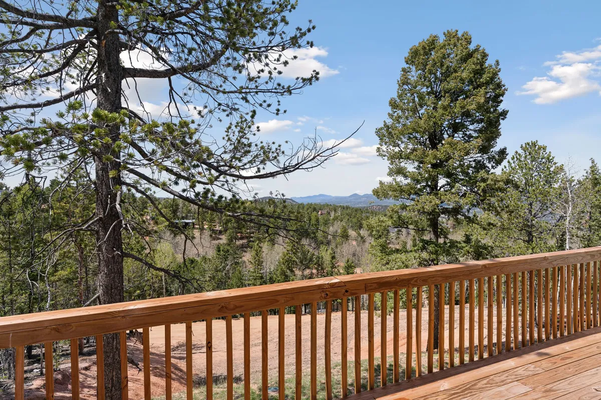 The image showcases a spacious wooden deck overlooking lush greenery, dramatic distant mountain peaks (including Pike's Peak), and a vast, open sky filled with soft clouds. Adirondack chairs are positioned to soak in the views, and the deck is designed for both relaxation and social gatherings.