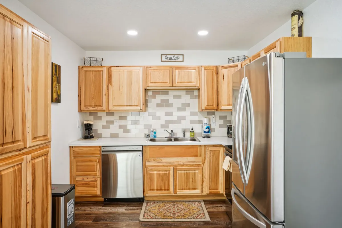 A cozy, functional kitchen corner with a prominent stainless steel refrigerator, wooden cabinets, a countertop with a sink, and a decorative rug. The space is designed for both practicality and social interaction.