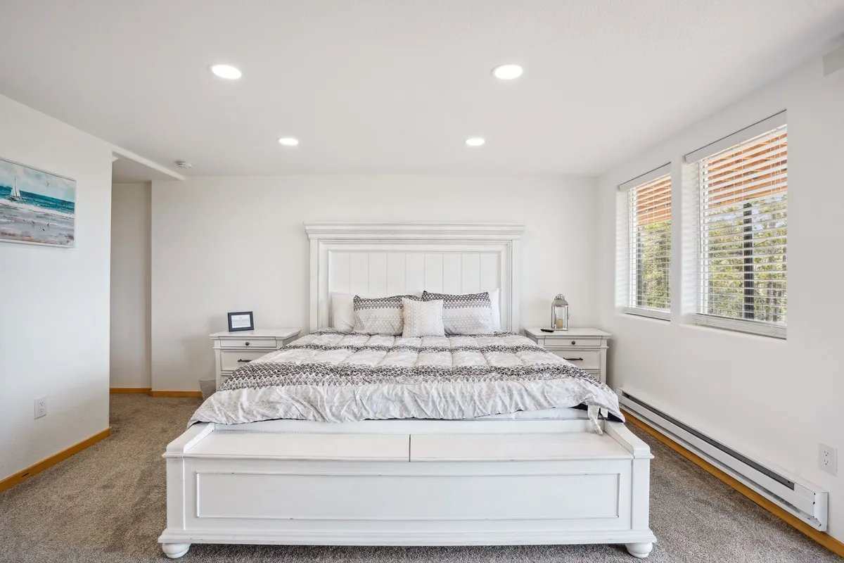 A serene bedroom with a large white bed adorned with decorative pillows, a side table with a lamp, and windows with blinds that softly filter in natural light. The overall environment feels restful and inviting, ideal for relaxation and quiet moments.