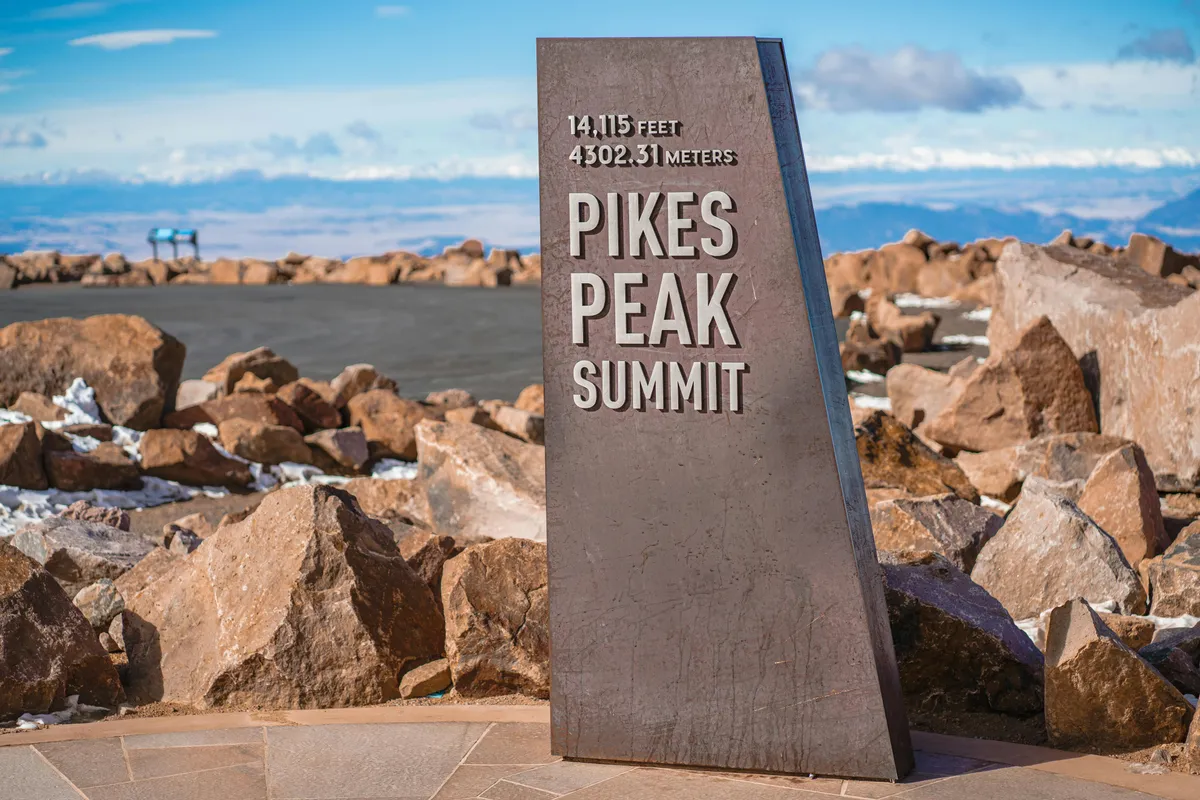 A triumphant scene at the summit of Pike's Peak, with a summit sign surrounded by unique rock formations and sweeping views of distant mountain ranges. The mood is adventurous, capturing the thrill of achievement after a challenging hike, and the panoramic vistas stretch for miles under Colorado’s open sky.