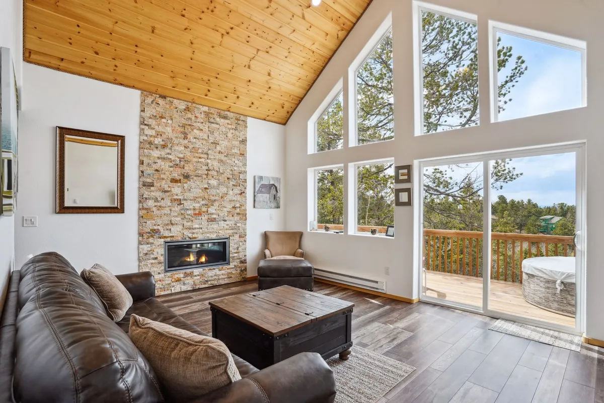 A spacious great room featuring a striking stone fireplace, wooden ceilings, a comfortable armchair, a large coffee table, and expansive windows that frame panoramic mountain views. Sunlight pours in, casting a warm, inviting glow throughout the space.