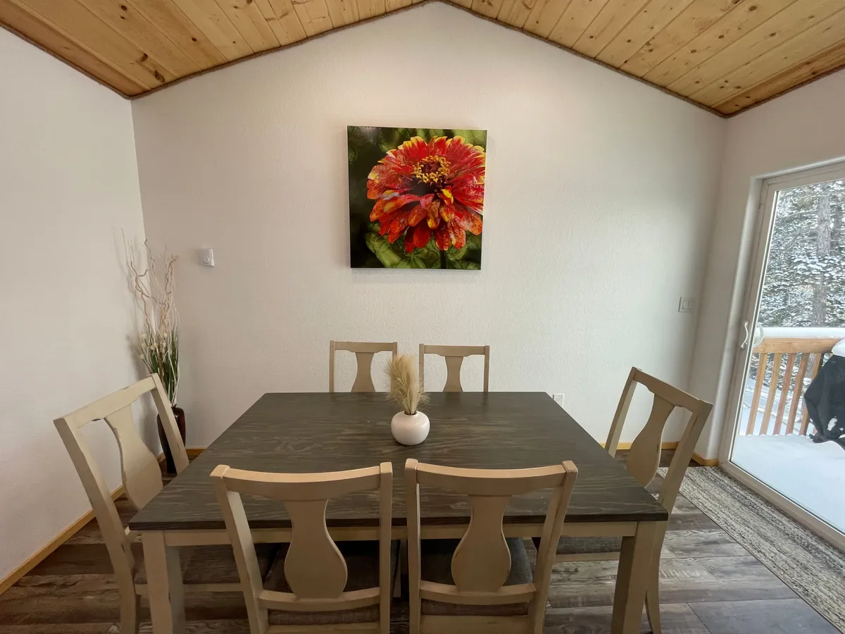 A welcoming dining area with a wooden table, floral artwork, a potted plant, and a window that frames an outdoor scene. The space is awash in morning sunlight, creating a cheerful, family-friendly ambiance.
