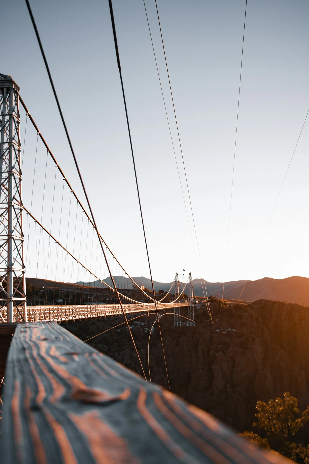 A striking suspension bridge stretches across an expansive mountain range under a vivid sunset sky. The scene captures the thrill of outdoor adventure and the awe-inspiring beauty of the surrounding environment, offering visual cues for exploration, discovery, and family-friendly activities.